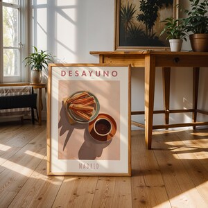May include: A framed art print featuring a breakfast scene with churros and coffee. The print has the word "DESAYUNO" at the top and "MADRID" at the bottom. The artwork is in a room with wooden floors and furniture.