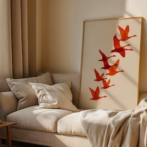 May include: A framed art print featuring a flock of orange geese in flight against a cream background. The artwork is displayed on a beige sofa with pillows and a blanket. The room has beige curtains and a wooden side table.