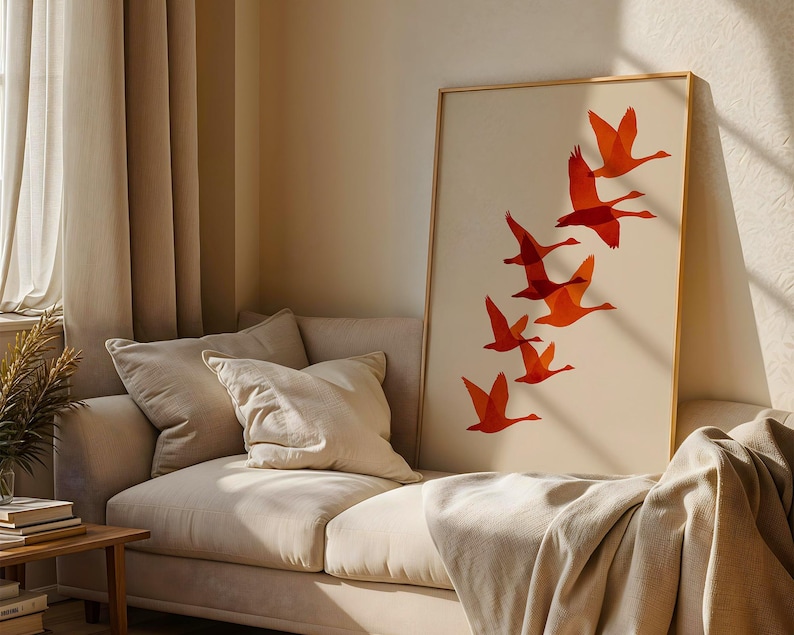 May include: A framed art print featuring a flock of orange geese in flight against a cream background. The artwork is displayed on a beige sofa with pillows and a blanket. The room has beige curtains and a wooden side table.