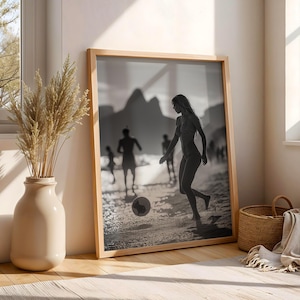 May include: Black and white framed print of people playing soccer on a beach. The image is in a light wooden frame, leaning against a wall. A beige vase with dried plants and a woven basket are in the foreground.