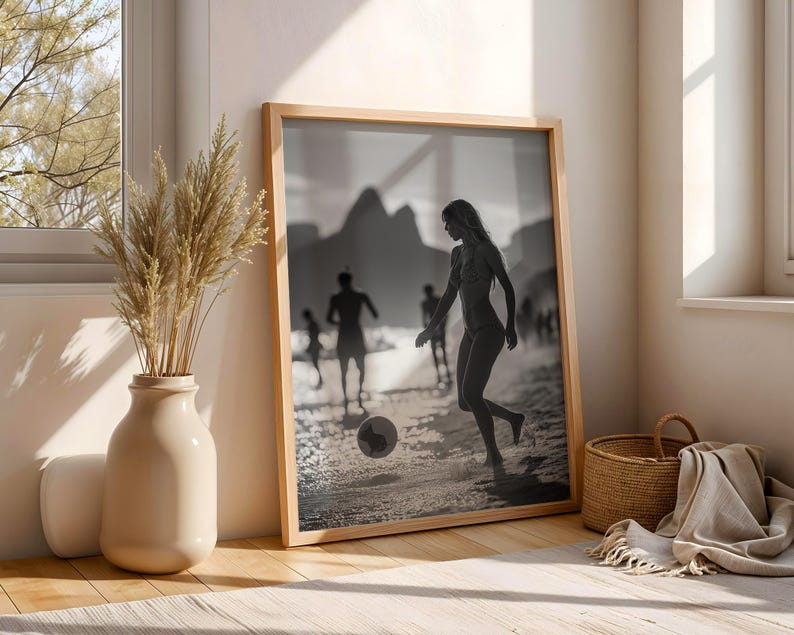 May include: Black and white framed print of people playing soccer on a beach. The image is in a light wooden frame, leaning against a wall. A beige vase with dried plants and a woven basket are in the foreground.