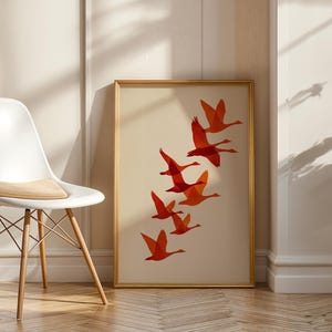 May include: A framed art print featuring a flock of orange birds in flight against a cream background. The artwork is displayed in a gold frame and leans against a wall. A white chair with wooden legs is in the foreground.