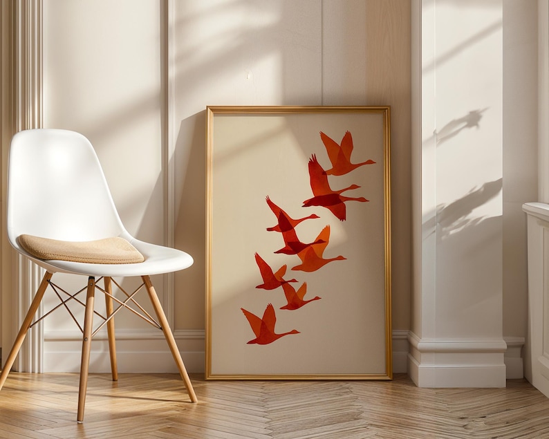 May include: A framed art print featuring a flock of orange birds in flight against a cream background. The artwork is displayed in a gold frame and leans against a wall. A white chair with wooden legs is in the foreground.