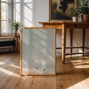 May include: A framed artwork with a light-colored background leans against a wall. The artwork features small figures and a red shape. The room has wooden floors, a wooden table, and a window with sunlight.