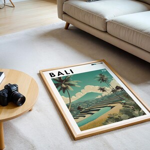 May include: A framed art print featuring a scenic landscape of Bali, Indonesia, with palm trees, terraced rice fields, and a mountain in the background. The word "BALI" is printed at the top. The frame is made of light-colored wood.