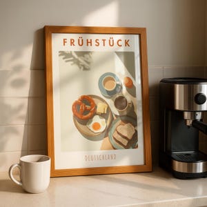 May include: A framed print with the word "FRÜHSTÜCK" and "DEUTSCHLAND" in orange text. The print depicts a breakfast scene with a pretzel, fried egg, coffee, and toast. A white mug and a coffee machine are on the counter.