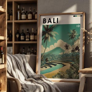 May include: A framed art print with the word "BALI" at the top. The print depicts a tropical landscape with palm trees, terraced fields, and a mountain under a teal sky. The print is in a wooden frame and is leaning against a wall.