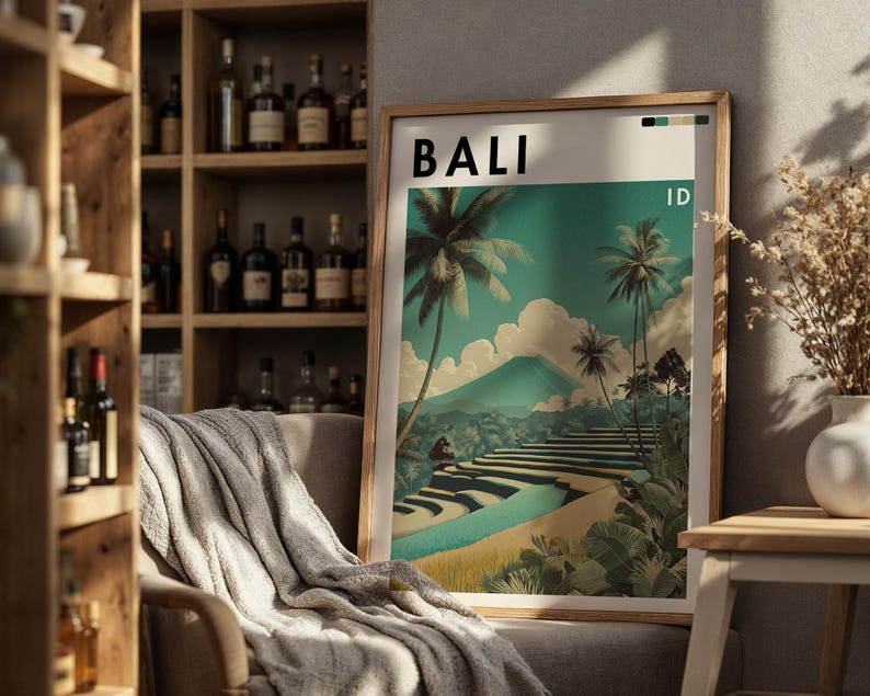 May include: A framed art print with the word "BALI" at the top. The print depicts a tropical landscape with palm trees, terraced fields, and a mountain under a teal sky. The print is in a wooden frame and is leaning against a wall.