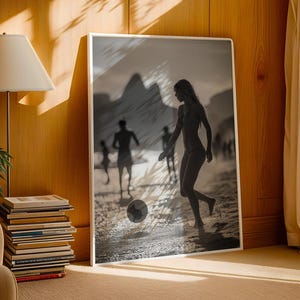 May include: Black and white framed print of a beach scene. A woman is kicking a soccer ball on the sand, with other figures in the background. The print is displayed in a room with a lamp, books, and a chair.