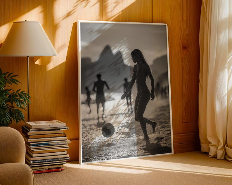 May include: Black and white framed print of a beach scene. A woman is kicking a soccer ball on the sand, with other figures in the background. The print is displayed in a room with a lamp, books, and a chair.