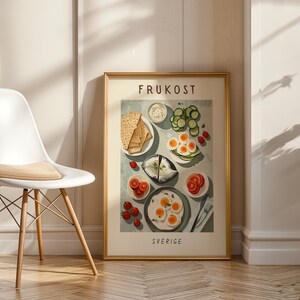 Puede incluir: Una impresión enmarcada que presenta una colorida ilustración de un desayuno sueco, que incluye huevos, tomates, pepinos y pan. La palabra "FRUKOST" está en la parte superior y "SVERIGE" en la parte inferior. La impresión está en un marco dorado.