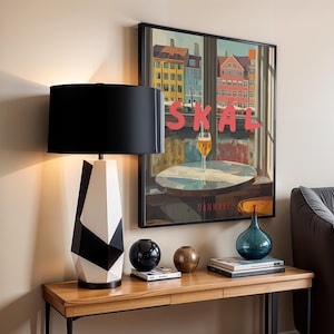 May include: A modern table lamp with a black lampshade and a geometric black and white base. The lamp sits on a wooden table with decorative items, including a blue glass vase and a framed poster with the word "SKAL".