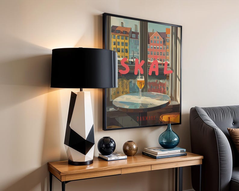 May include: A modern table lamp with a black lampshade and a geometric black and white base. The lamp sits on a wooden table with decorative items, including a blue glass vase and a framed poster with the word "SKAL".