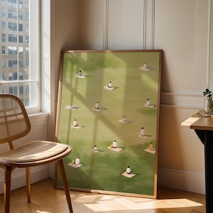May include: A framed art print featuring a green field with small figures on mats. The artwork is in a wooden frame and leans against a wall next to a window and a wooden chair. The room has hardwood floors.