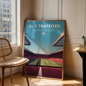 May include: A framed art print of Old Trafford stadium in Manchester, England. The print features a stylized view of the stadium with a blue sky, red seating, and green field. The text "OLD TRAFFORD MANCHESTER" is at the top.
