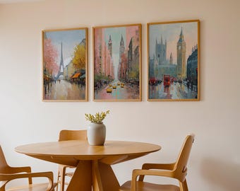 May include: Three framed city paintings hang above a wooden table and chairs. The paintings depict scenes of Paris, New York, and London. The artwork features a color palette of pinks, yellows, and blues. A vase with greenery sits on the table.