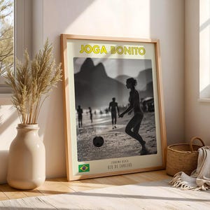 Może przedstawiać: Oprawiony plakat ze słowami "Joga Bonito" w ż&oacute;łtych literach. Czarno-biały obraz przedstawia osobę grającą w piłkę nożną na plaży. Brazylijska flaga znajduje się w lewym dolnym rogu, z tekstem "Ipanema Beach, Rio de Janeiro."