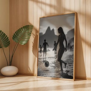 May include: A framed black and white print depicts a person playing soccer on a beach. The image is set in a wooden frame and leans against a wall. A potted plant is in the foreground.
