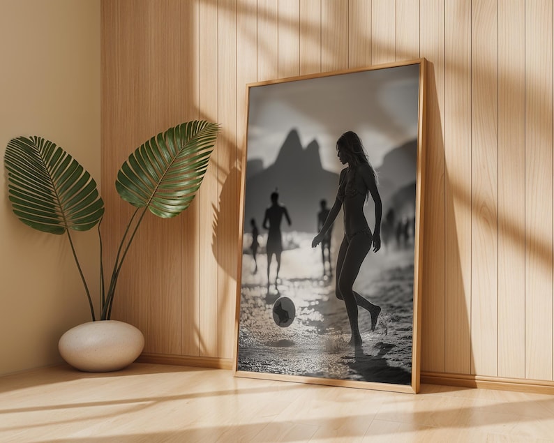 May include: A framed black and white print depicts a person playing soccer on a beach. The image is set in a wooden frame and leans against a wall. A potted plant is in the foreground.