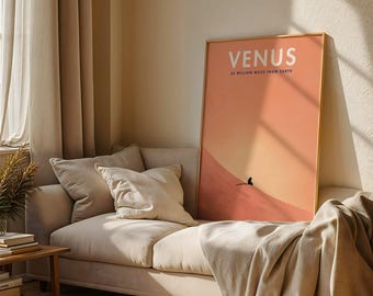 May include: A framed art print with the word "VENUS" in white text, and the phrase "25 MILLION MILES FROM EARTH" below it. The print has a gradient of orange and peach colors. The print is displayed on a beige sofa with pillows and a blanket.