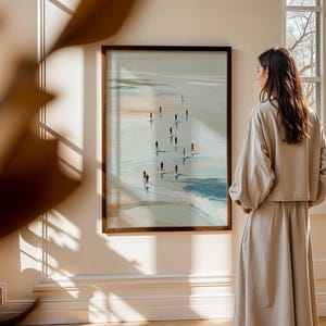May include: A framed artwork depicts figures on surfboards in a coastal scene, with a palette of blues, greens, and sandy tones. The artwork is displayed on a cream-colored wall, and a person is standing in front of the artwork. The artwork is in a room with natural light.