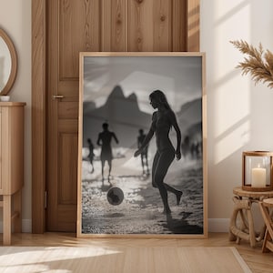 May include: Black and white framed print of a person playing soccer on a beach. The image features a woman kicking a soccer ball, with other people in the background. The print is displayed in a light wood frame, leaning against a wooden door.