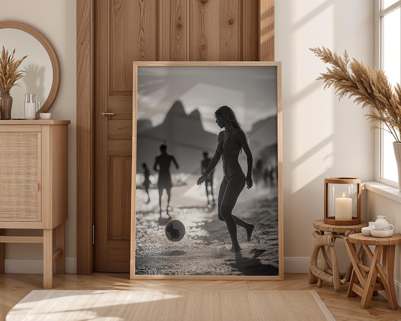May include: Black and white framed print of a person playing soccer on a beach. The image features a woman kicking a soccer ball, with other people in the background. The print is displayed in a light wood frame, leaning against a wooden door.