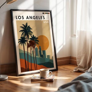 May include: A framed art print featuring a Los Angeles landscape with palm trees, a setting sun, and city skyline. The print has a black frame and is leaning against a wall. A coffee cup and book are on the floor in front of the print.