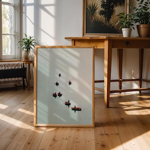 Op de afbeelding: Een ingelijste kunstdruk met kleine boten met mensen op een lichtblauwe achtergrond. Het kunstwerk wordt op een houten vloer tentoongesteld, leunend tegen een muur. De kamer heeft natuurlijk licht en houten meubels.