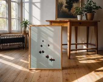 May include: A framed art print featuring small boats with people on a light blue background. The artwork is displayed on a wooden floor, leaning against a wall. The room has natural light and wooden furniture.