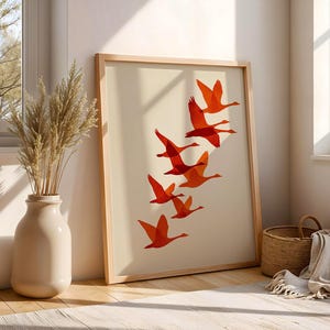 May include: A framed art print featuring a flock of orange geese in flight against a cream background. The artwork is displayed in a light wood frame, leaning against a wall. A vase with dried plants and a woven basket are nearby.