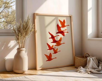 May include: A framed art print featuring a flock of orange geese in flight against a cream background. The artwork is displayed in a light wood frame, leaning against a wall. A vase with dried plants and a woven basket are nearby.