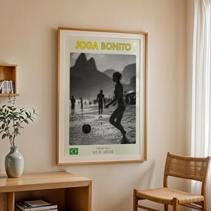 Może przedstawiać: Oprawiona czarno-biała fotografia osoby grającej w piłkę nożną na plaży. Obraz zawiera ż&oacute;łty napis "Joga Bonito" i brazylijską flagę. Fotografia jest w drewnianej ramie.
