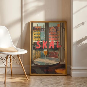 May include: A framed art print featuring a colorful cityscape with the word "SKAL" in large pink letters. A glass of beer sits on a table in front of the window. The print is displayed next to a white chair.