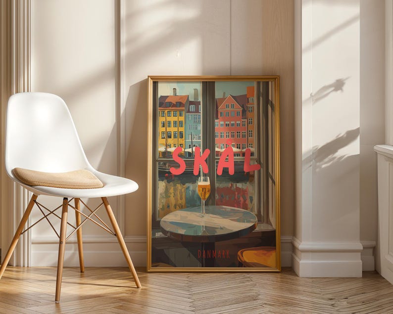 May include: A framed art print featuring a colorful cityscape with the word "SKAL" in large pink letters. A glass of beer sits on a table in front of the window. The print is displayed next to a white chair.
