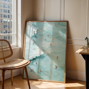 May include: A framed art print depicts a serene scene of people swimming in a turquoise body of water. The artwork, with a light wood frame, is leaning against a wall next to a window and a wooden chair. The room is bathed in natural light.