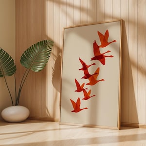 May include: A framed art print featuring a flock of orange geese in flight against a cream background. The artwork is displayed in a light wood frame, leaning against a wooden wall. A potted plant with large green leaves is in the foreground.