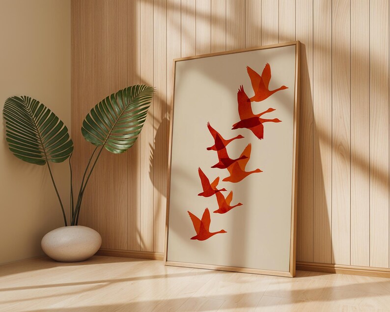 May include: A framed art print featuring a flock of orange geese in flight against a cream background. The artwork is displayed in a light wood frame, leaning against a wooden wall. A potted plant with large green leaves is in the foreground.