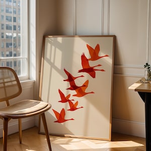 May include: A framed art print featuring a flock of orange geese in flight against a cream background. The artwork is displayed in a room with a wooden chair, a window, and a small table.