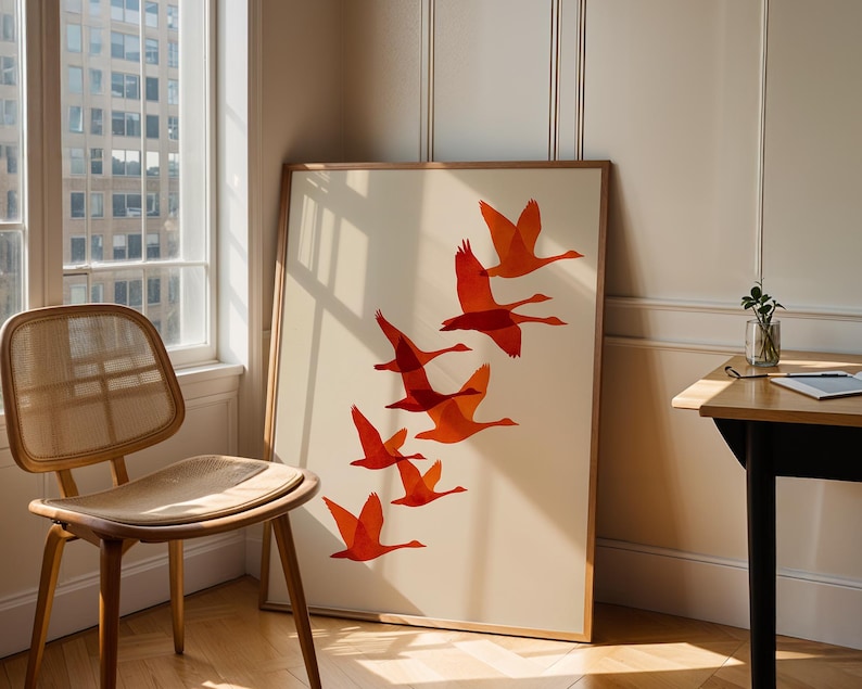 May include: A framed art print featuring a flock of orange geese in flight against a cream background. The artwork is displayed in a room with a wooden chair, a window, and a small table.
