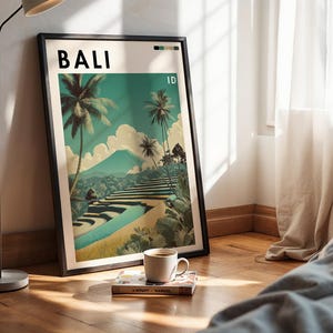 May include: A framed art print featuring a Bali landscape with palm trees, terraced rice fields, and a mountain. The print has the word "BALI" at the top. A coffee cup and a book rest on the wooden floor in front of the print.