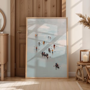 Op de afbeelding: Een ingelijste kunstdruk toont verschillende kleine boten met mensen die op een watermassa roeien. Het kunstwerk heeft een lichtblauw en wit kleurenpalet, met een houten lijst. De print wordt tentoongesteld in een goed verlichte kamer met houten meubels.