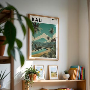 May include: A framed poster with the word "BALI" at the top, showcasing a scenic landscape with palm trees, terraced fields, and a mountain. The poster is in a light wood frame and hangs on a white wall. Other framed pictures and plants are on a wooden shelf below.