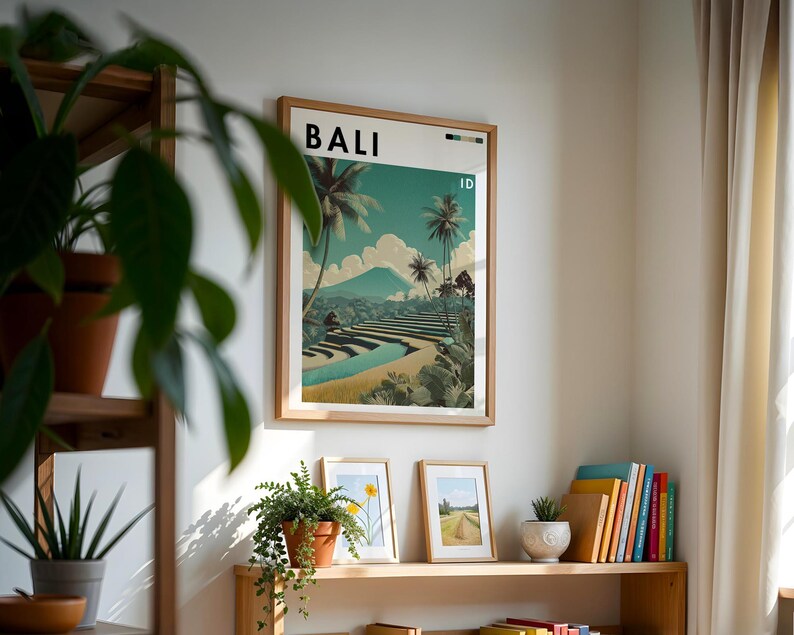 May include: A framed poster with the word "BALI" at the top, showcasing a scenic landscape with palm trees, terraced fields, and a mountain. The poster is in a light wood frame and hangs on a white wall. Other framed pictures and plants are on a wooden shelf below.