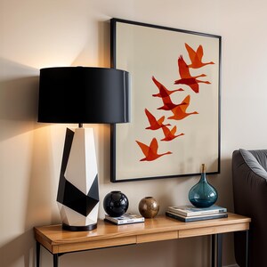 May include: A modern table lamp with a black lampshade and a geometric black and white base. A framed artwork with orange birds in flight hangs on the wall. Decorative spheres and a blue glass bottle sit on a wooden table.