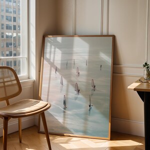 May include: A framed art print featuring windsurfers on a body of water. The artwork has a light wood frame and is leaning against a wall next to a wooden chair with a woven seat and back. A small table with a plant and a window are also in the scene.