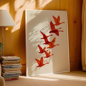 May include: A framed art print featuring a flock of orange birds in flight against a cream background. The artwork is displayed in a white frame and leans against a wooden wall. The room is lit by natural light.