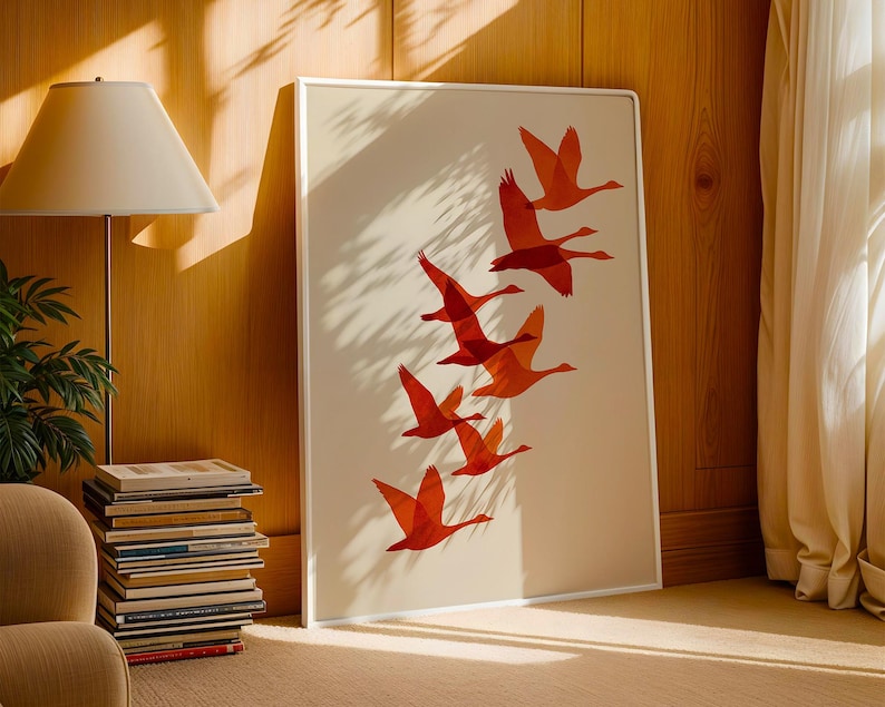 May include: A framed art print featuring a flock of orange birds in flight against a cream background. The artwork is displayed in a white frame and leans against a wooden wall. The room is lit by natural light.