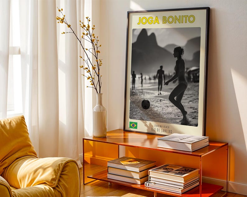 Może przedstawiać: Oprawiony plakat z ż&oacute;łtym napisem "Joga Bonito", przedstawiający czarno-biały obraz osoby grającej w piłkę nożną na plaży. Plakat jest umieszczony na pomarańczowym stoliku z książkami i wazonem z ż&oacute;łtymi kwiatami.