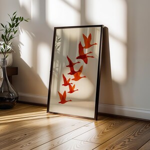 May include: A framed art print featuring a flock of orange birds in flight against a cream background. The artwork is displayed in a dark brown frame and leans against a white wall. The room has hardwood floors and a glass vase with greenery.